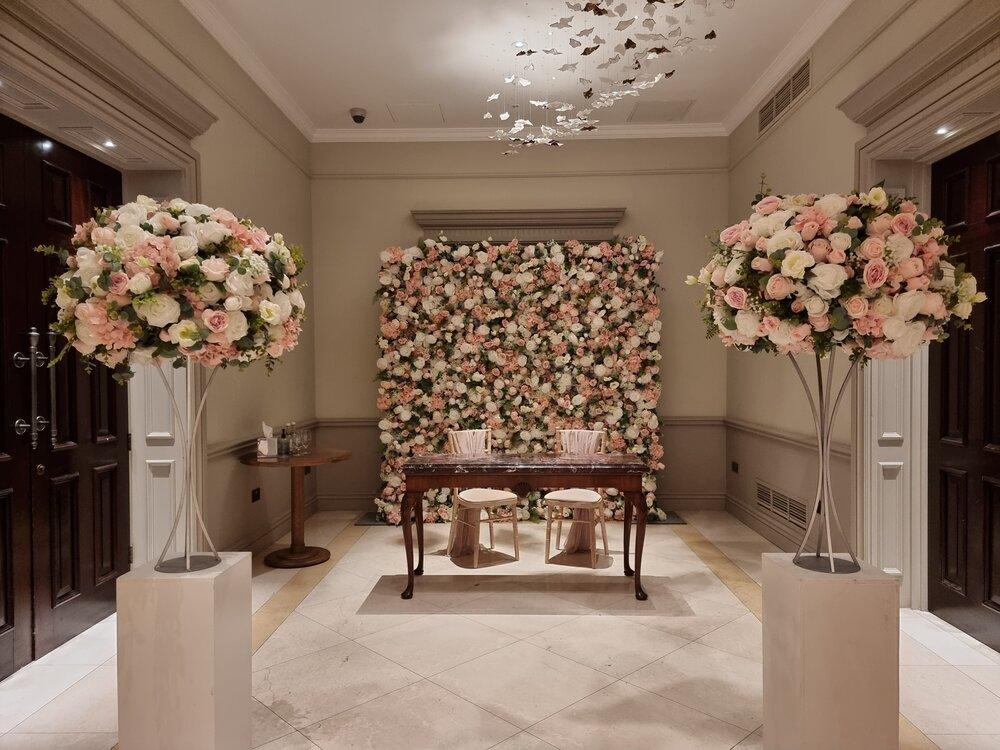 a floral wall with a table in front of it and two matching bouquets | Wedifys