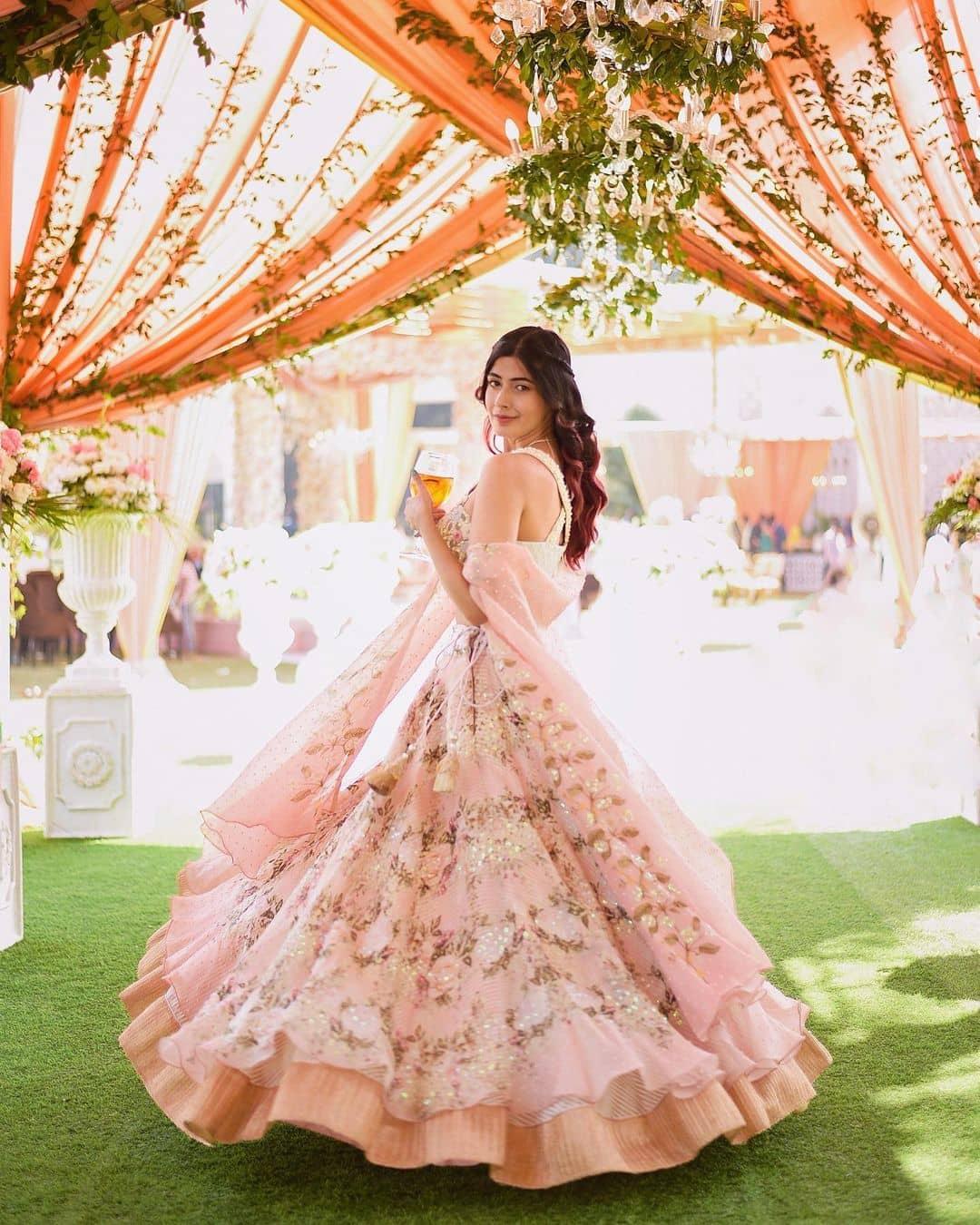 a woman wearing a baby pink floral lehenga while holding a glass of drink in her hand | Wedifys