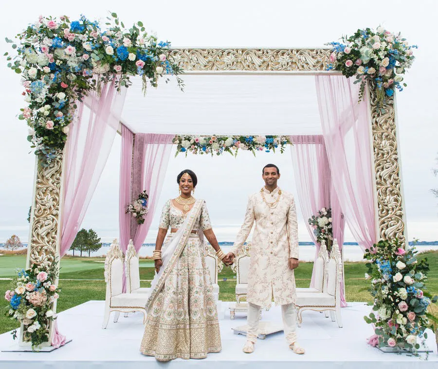 bride and groom standing in a sleek gold mandap ornated with blue and pink flowers | Wedifys