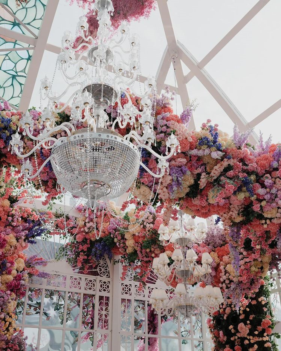 a beautiful chandelier can be seen with multicolor floral decorations around the ceiling and pillars. | Anant Ambani and Radhika Merchant Pre-Wedding Decor | Wedifys